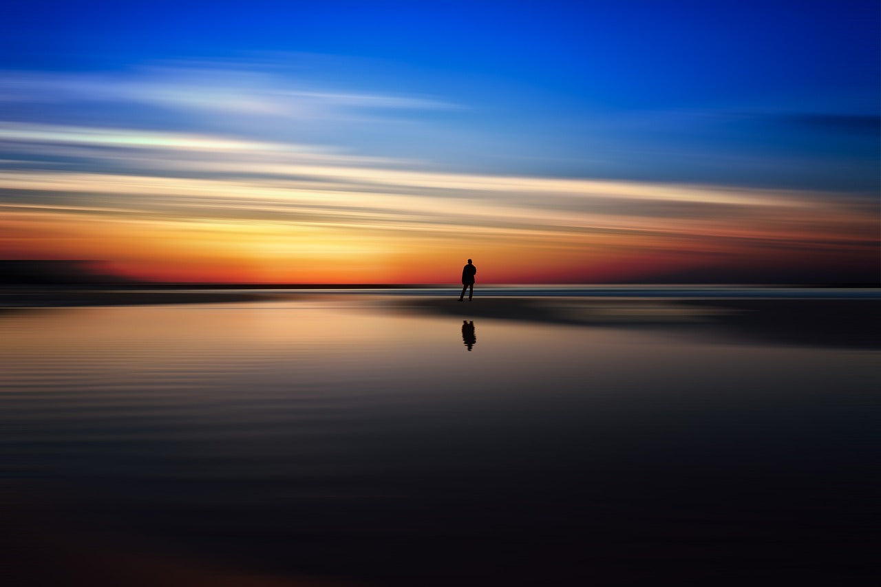 A lone figure standing on a beach at sunset, with colorful clouds reflected on the water surface.