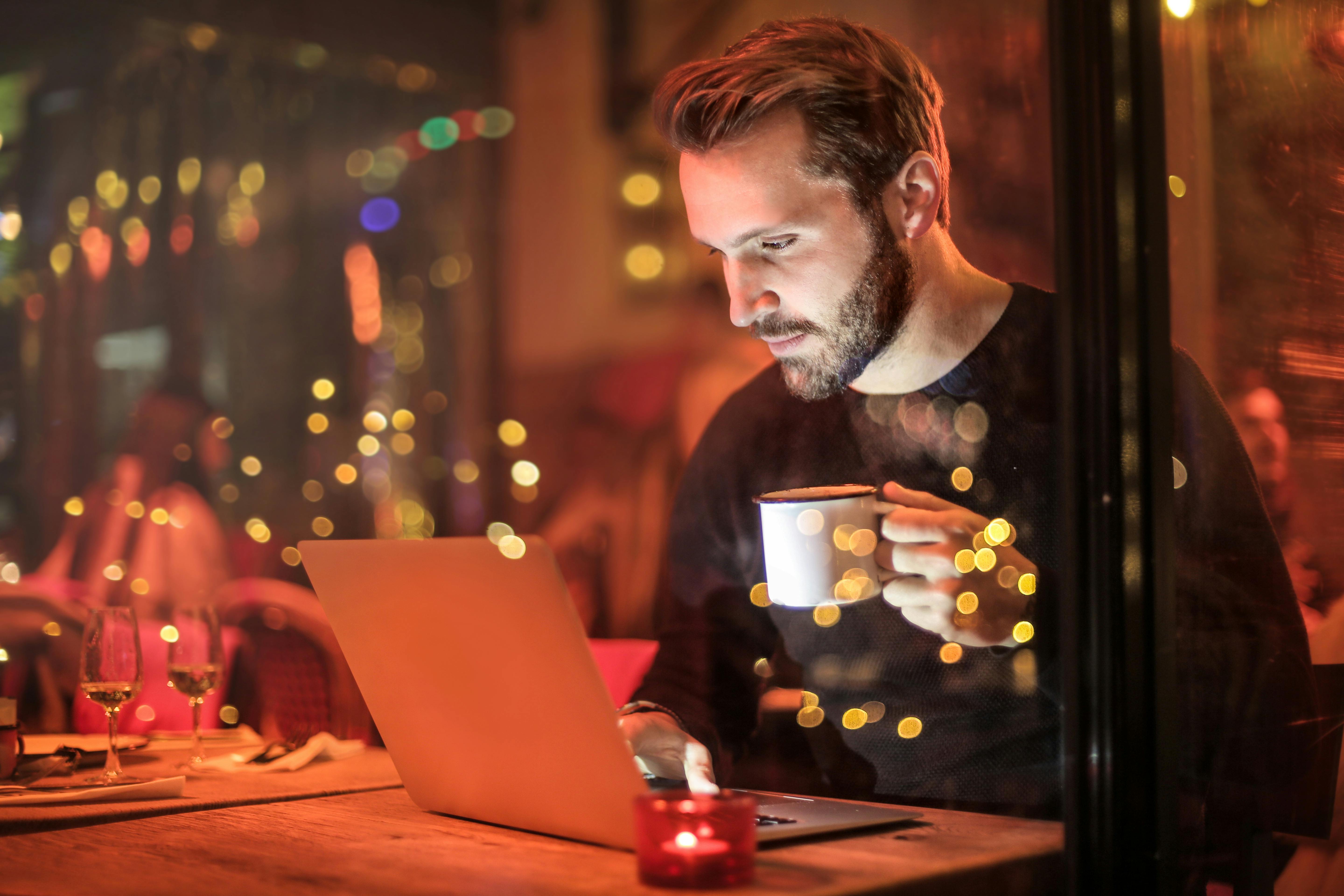 Man working on a laptop in a cozy café at night, holding a coffee cup, with warm bokeh lights in the background.