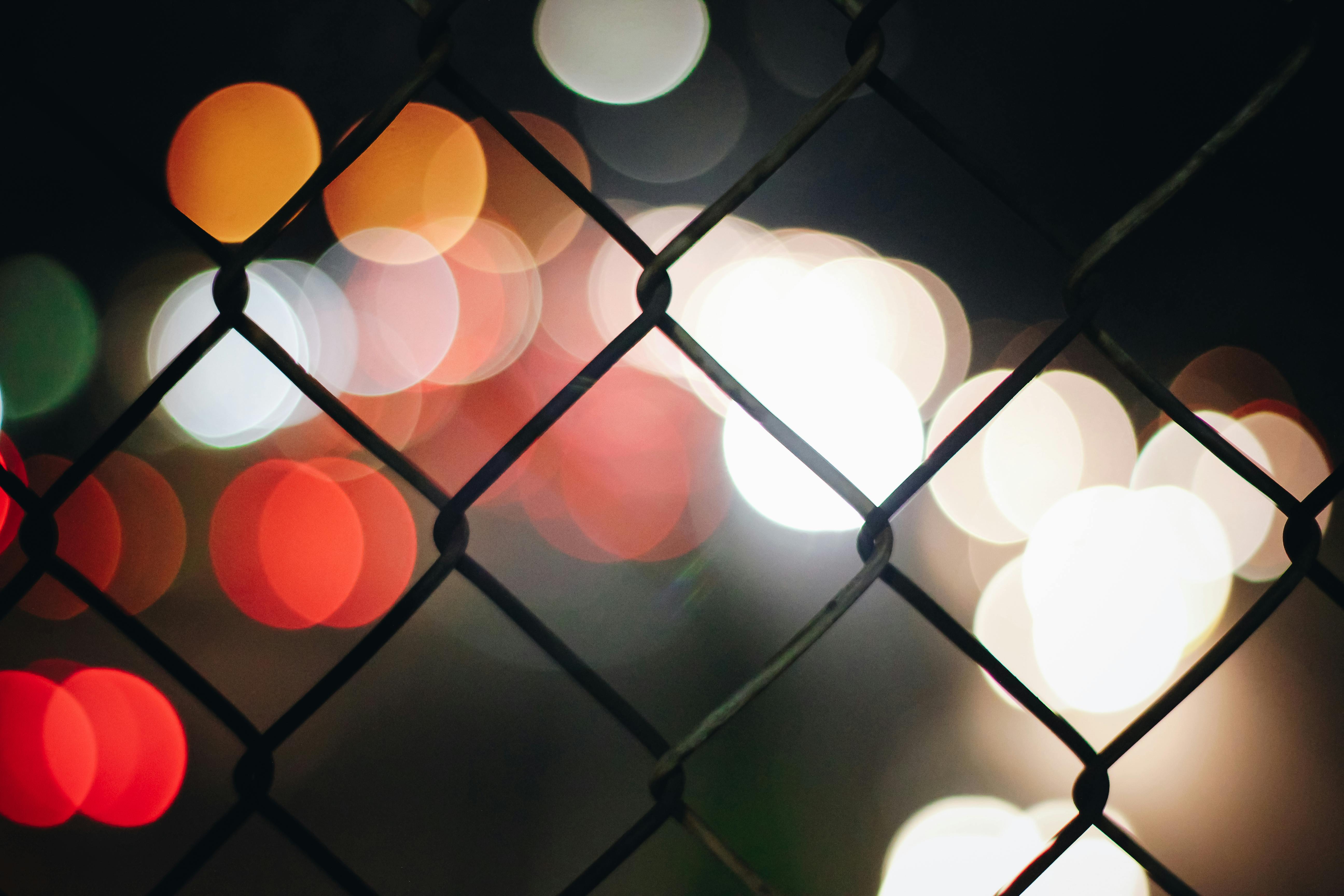 Bokeh lights behind a chain-link fence at night, creating a colorful and abstract effect.